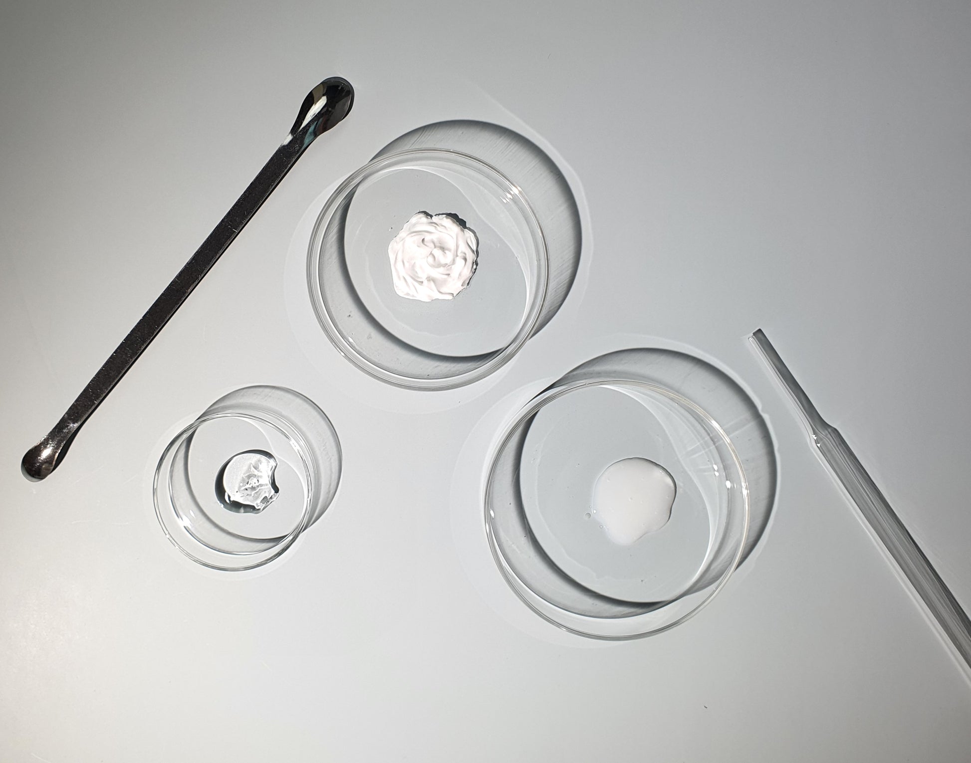 Three petri dishes with skin essence, moisturizer and gel inside. Spatula and dropper lying on both sides.