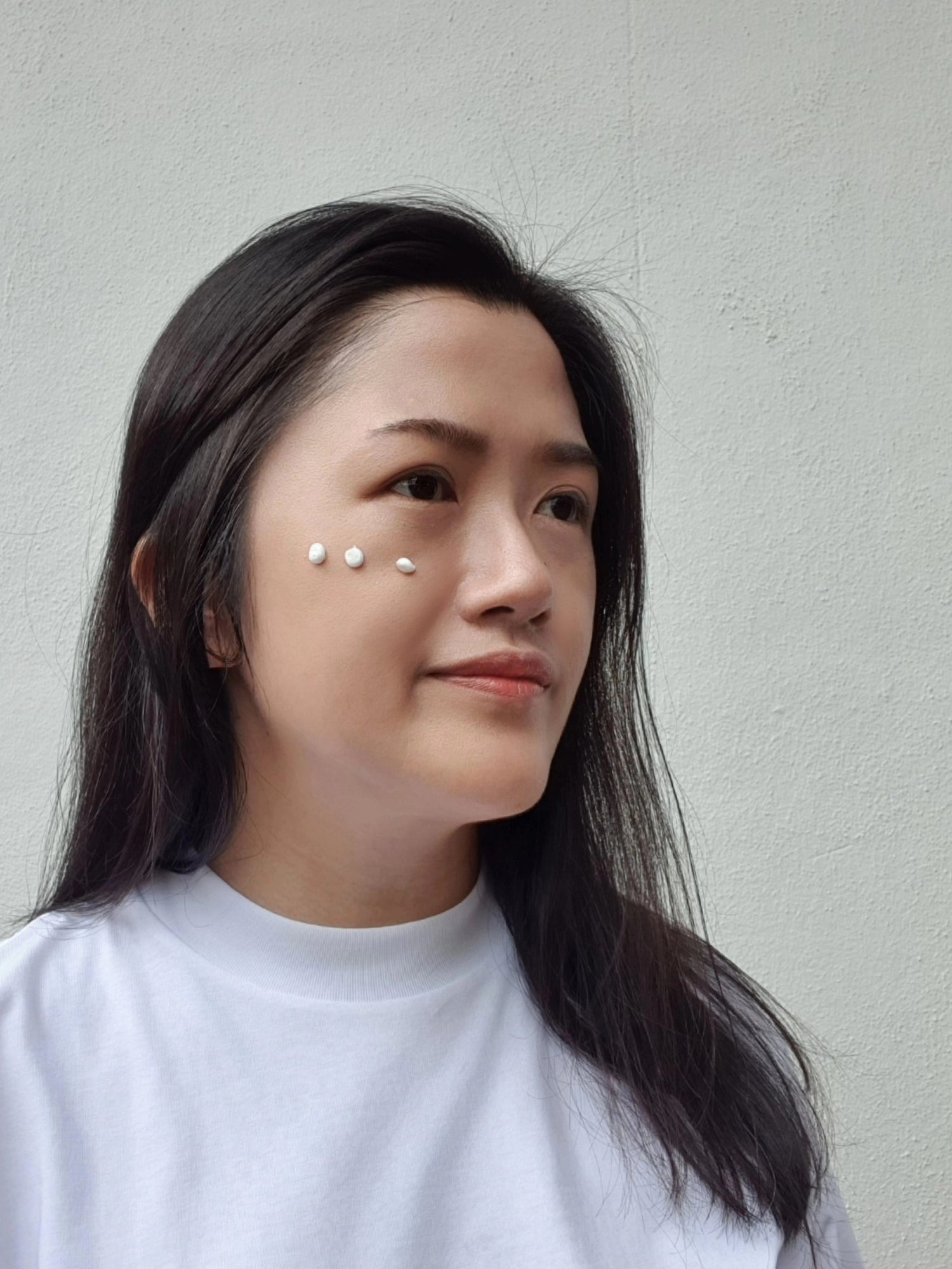 Smiling woman with three dots of moisturizer on her cheek against a white background.