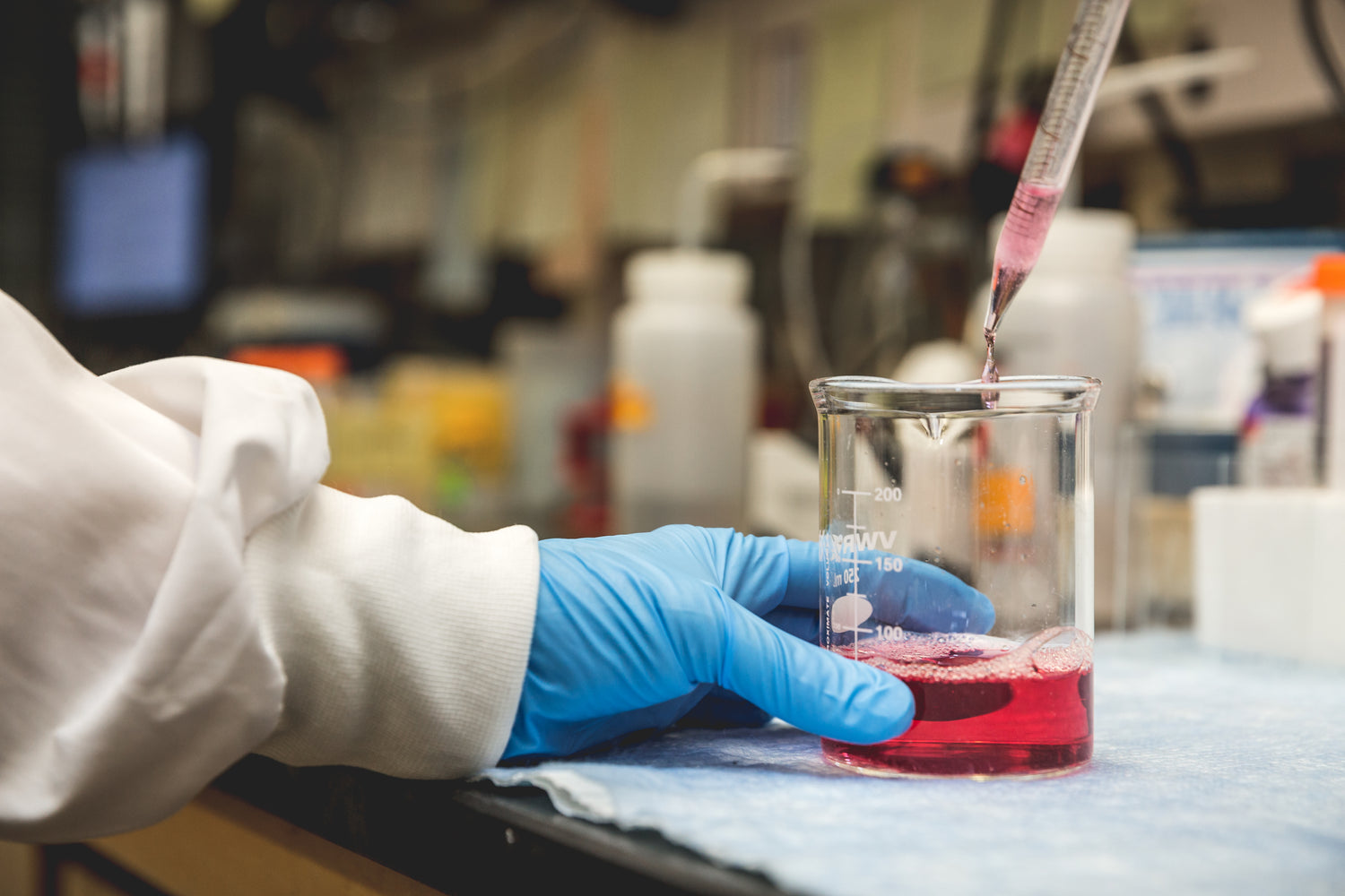 Chemist wearing blue gloves holding a beaker filled with red transparent liquid.